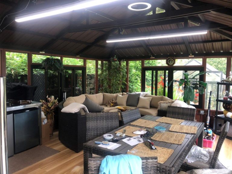 Spacious sunroom with wicker furniture, a large sectional couch, plants, and windows overlooking lush greenery.