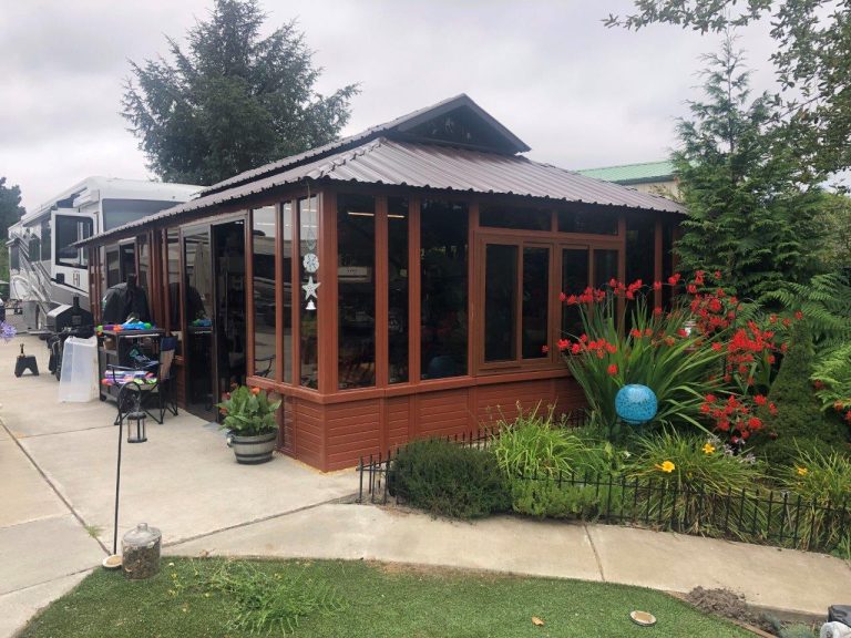 A wooden garden shed with large windows, surrounded by red flowers and greenery on a cloudy day.