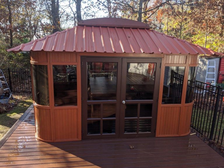 Wooden gazebo with a red metal roof on a deck, surrounded by trees and a black fence.