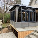 Gray sunroom with large windows on a wooden deck, surrounded by trees and a stone face sculpture nearby.