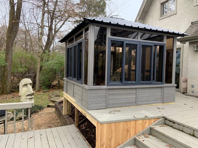 Gray sunroom with large windows on a wooden deck, surrounded by trees and a stone face sculpture nearby.