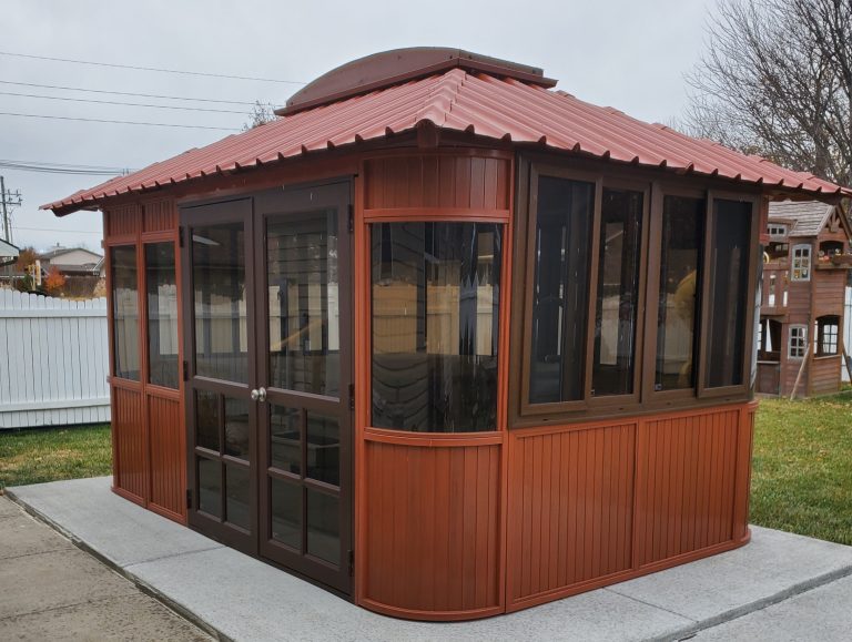 A gazebo with a red metal roof and glass windows, set on a concrete pad in a backyard.