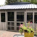 A modern gazebo with glass windows and a metal roof, surrounded by potted plants and trees.