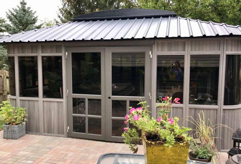 A modern gazebo with glass windows and a metal roof, surrounded by potted plants and trees.
