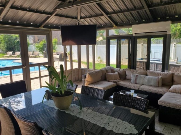 Covered patio with wicker furniture, dining table, TV, and pool view through glass doors.