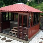 The Aspen 12x12, a red-roofed wooden gazebo with glass doors and windows, sits on a stone patio with white rocks and wooden steps—ideal as a stylish swim spa enclosure surrounded by lush greenery.