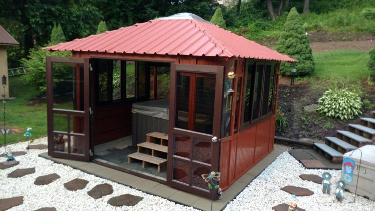 The Aspen 12x12, a red-roofed wooden gazebo with glass doors and windows, sits on a stone patio with white rocks and wooden steps—ideal as a stylish swim spa enclosure surrounded by lush greenery.