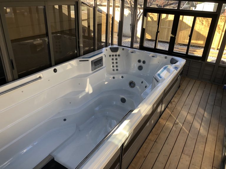 An empty indoor swim spa with a wooden deck and large windows surrounding it.