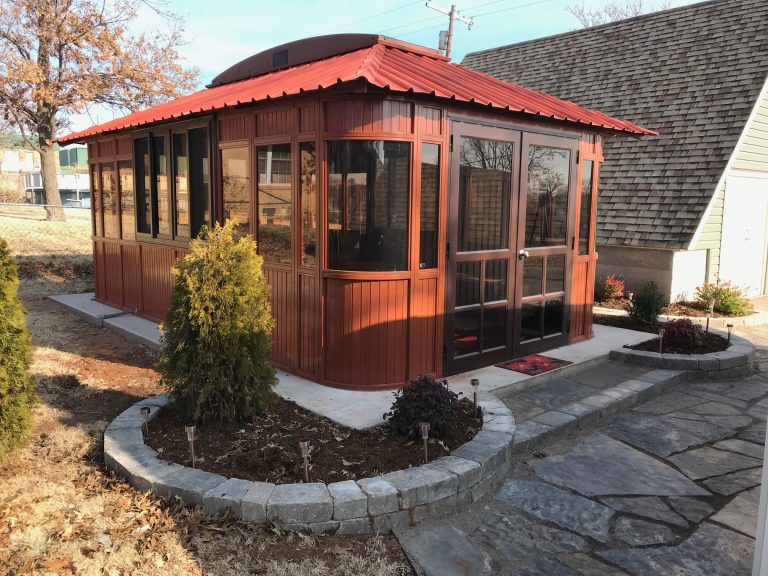 A red-roofed wooden gazebo with large windows sits next to a house, surrounded by small bushes and stone pavement.