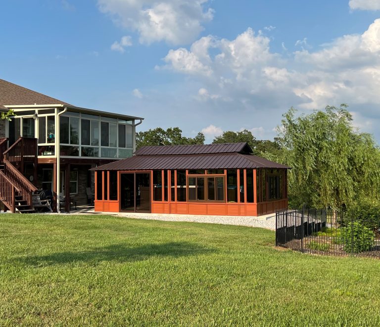 A modern sunroom with large windows next to a wooden house, surrounded by greenery and a clear blue sky.