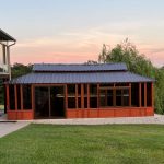 Red wooden gazebo with a blue metal roof and glass panels, set in a grassy area near trees at sunset.