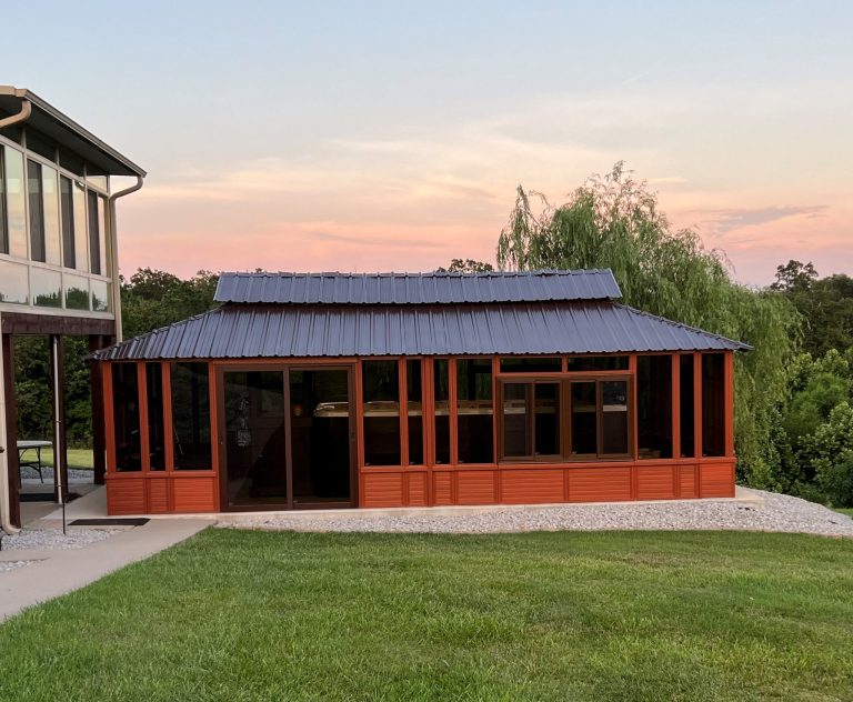 Red wooden gazebo with a blue metal roof and glass panels, set in a grassy area near trees at sunset.