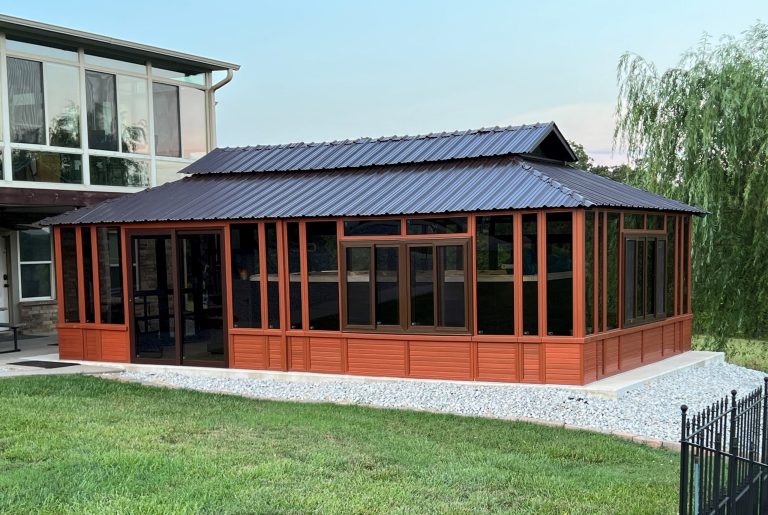 Modern gazebo with large windows and a corrugated metal roof, beside a house, surrounded by grass.