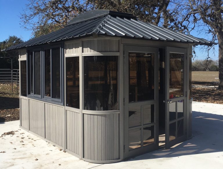 Gray gazebo with large windows and a corrugated metal roof set on a concrete slab in a park.