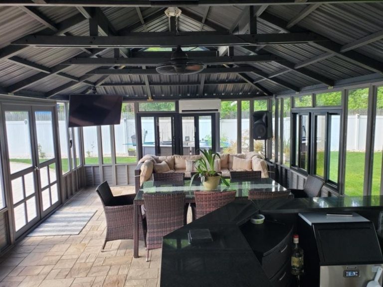 Enclosed patio with glass walls, wicker furniture, a TV, ceiling fan, and greenery outside; modern and cozy setting.