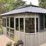 A gray wooden gazebo with glass windows and a metal roof, surrounded by trees and potted plants.