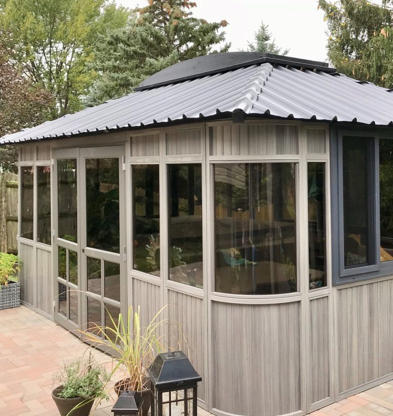 A gray wooden gazebo with glass windows and a metal roof, surrounded by trees and potted plants.