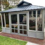 A small gray gazebo with a metal roof and glass doors, surrounded by grass and trees.