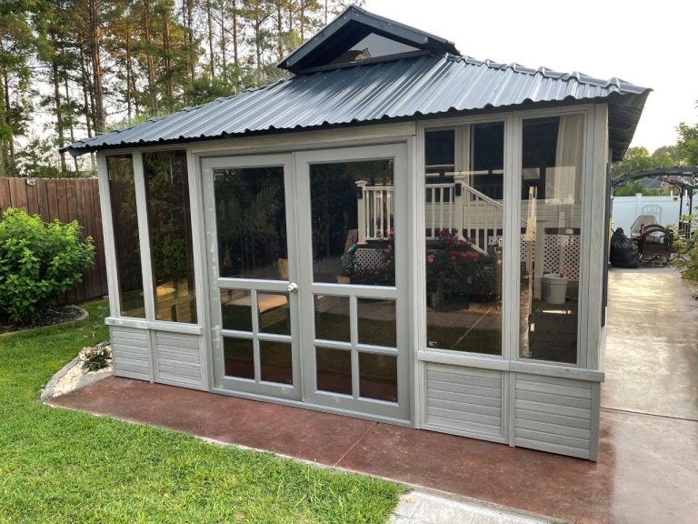 A small gray gazebo with a metal roof and glass doors, surrounded by grass and trees.