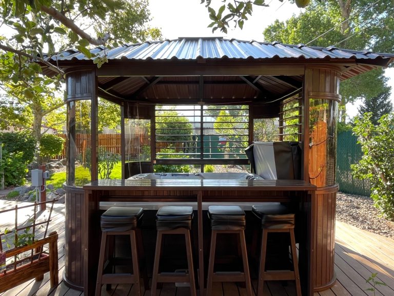 A wooden gazebo bar setup with stools and a covered grill, surrounded by a garden with a wooden fence.
