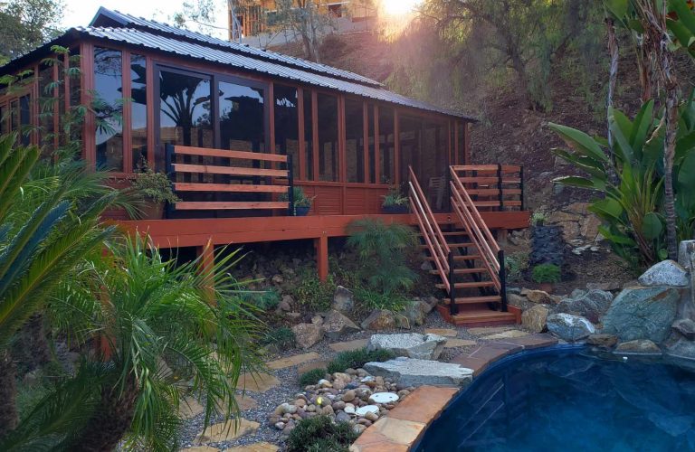 Wooden terrace house with stairs, surrounded by plants and rocks, near a blue pool.