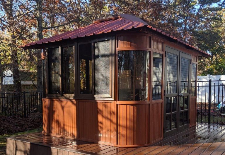 A small brown wooden gazebo with windows and a red roof stands on a wooden deck, surrounded by trees.
