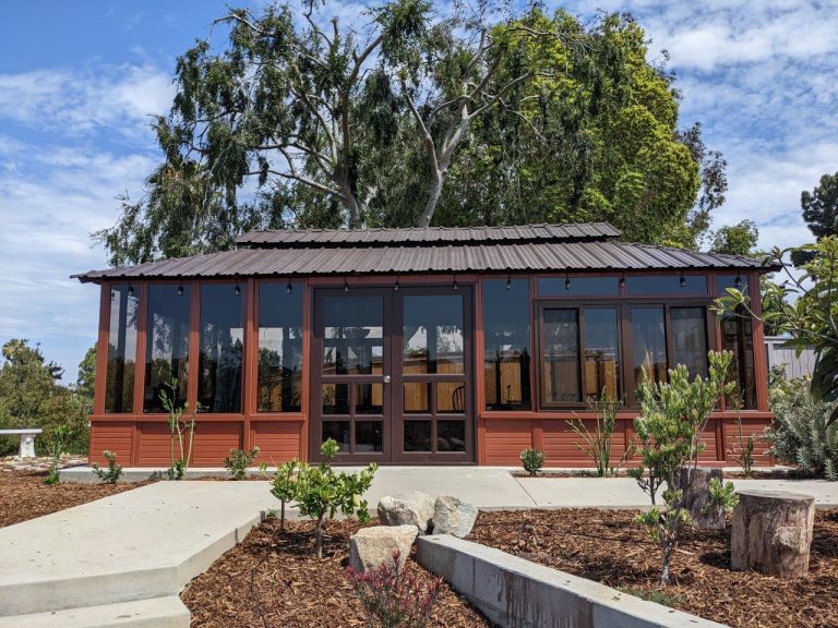Wooden garden shed with a metal roof and large windows, surrounded by greenery and a clear sky.