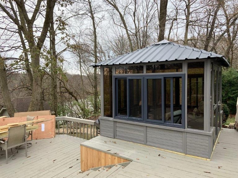 Gray gazebo on a wooden deck surrounded by bare trees, with outdoor furniture and construction materials nearby.