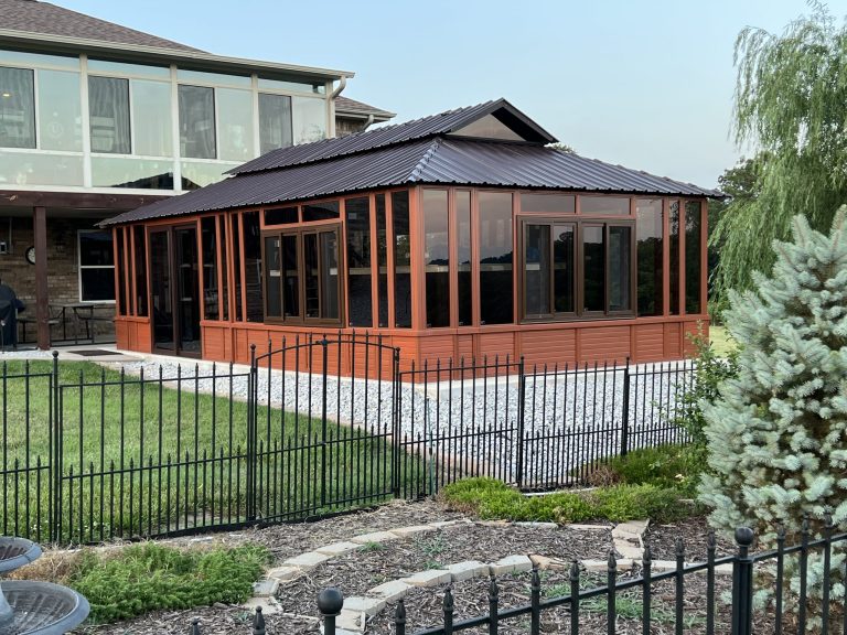 Red gazebo with dark roof beside a large house, surrounded by a black fence, greenery, and a garden path.