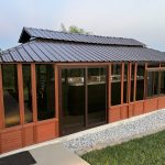 Brown gazebo with glass panels and a metal roof surrounded by grass and a rock border.