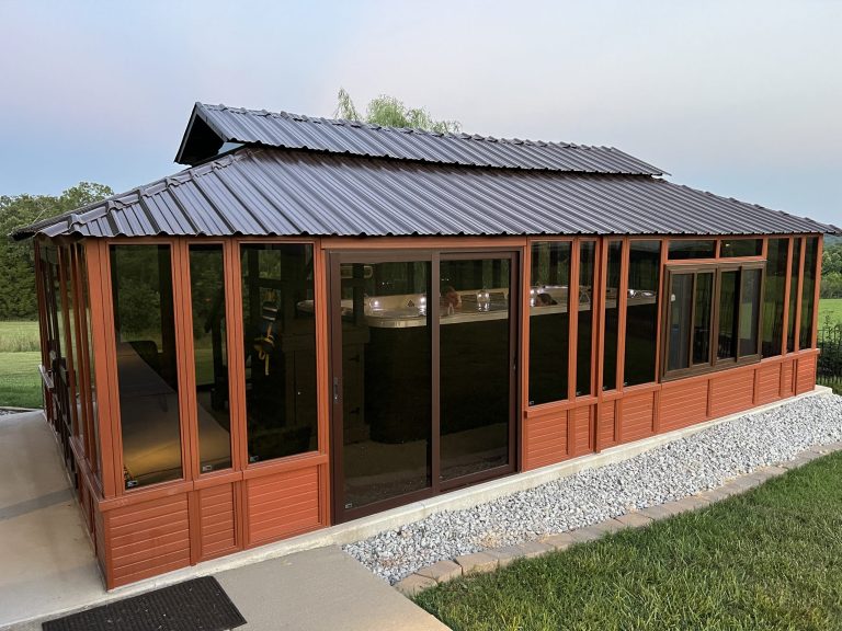 Brown gazebo with glass panels and a metal roof surrounded by grass and a rock border.