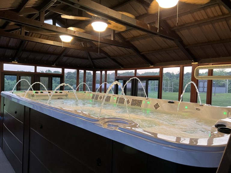 Indoor whirlpool with water jets under a wooden roof, surrounded by windows and a view of greenery outside.