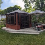 Garden with a gazebo, striped umbrella, metal chairs, and a red bench on a concrete patio surrounded by greenery.