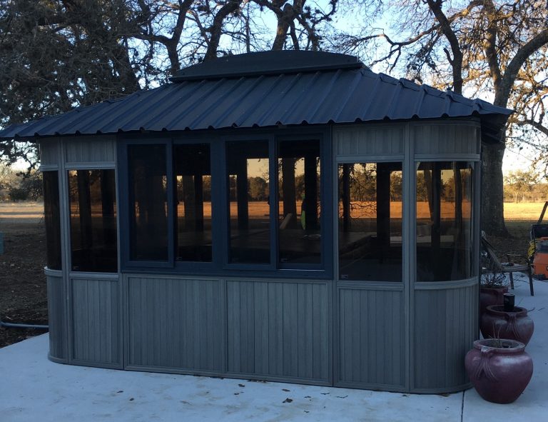 Gray gazebo with dark windows on concrete surrounded by trees and red planters.