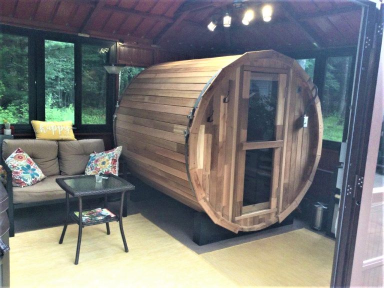 Wooden barrel sauna in a cozy room with a sofa, colorful pillows, and small table.