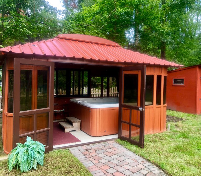 Wooden gazebo with a red metal roof, housing a hot tub, set in a grassy backyard with trees.
