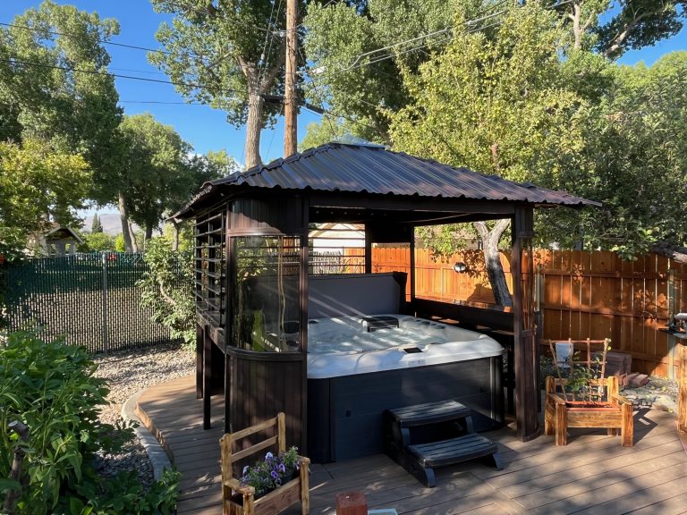 Gazebo with a hot tub inside, surrounded by trees and a fence, on a sunny day.