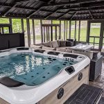 Indoor gazebo with a large hot tub, surrounded by wicker furniture and glass doors.