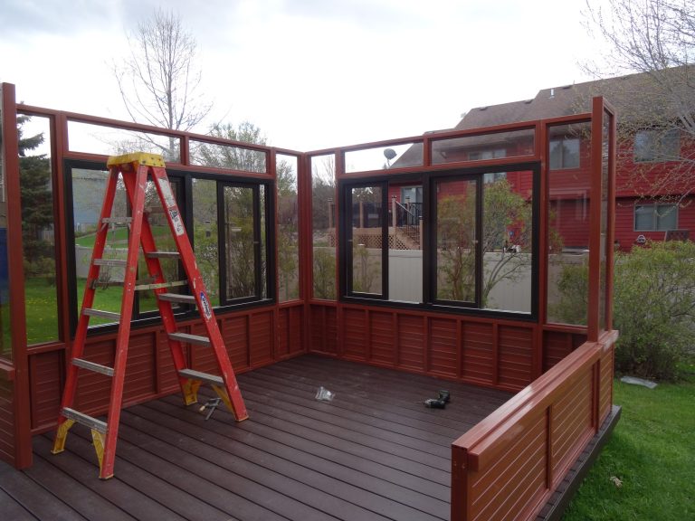 Red ladder inside an unfinished outdoor structure with windows, on a deck, with houses and trees in the background.