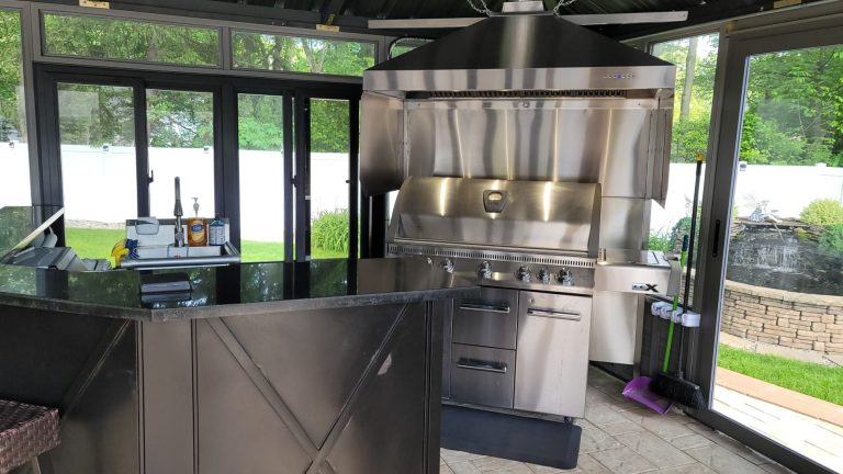 Outdoor kitchen with a large stainless steel grill, black counter, and large windows overlooking greenery.