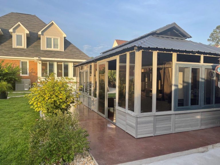 A patio with a screened gazebo next to a two-story house, surrounded by green lawn and bushes on a sunny day.