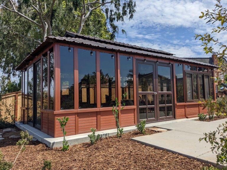 A red wooden greenhouse with large glass windows, surrounded by trees and a clear sky.