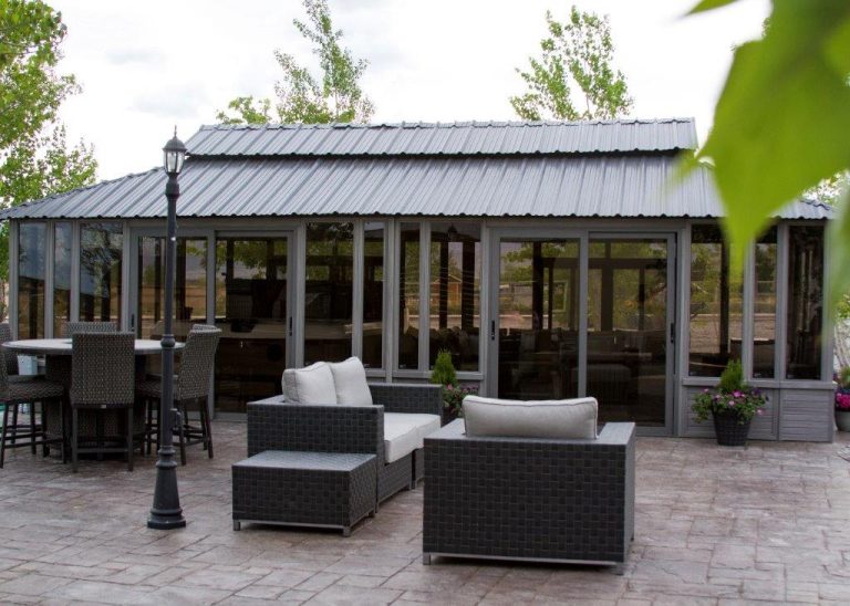 Outdoor patio with wicker furniture, a metal-roofed sunroom, trees, and potted plants on a stone terrace.