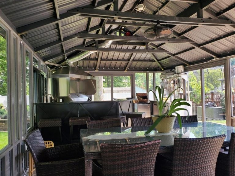 Outdoor kitchen with metal roof, glass table, wicker chairs, and potted plant under a fan, surrounded by greenery.