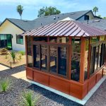 A small glass-and-wood gazebo with a metal roof in a landscaped backyard with desert plants and a modern house.