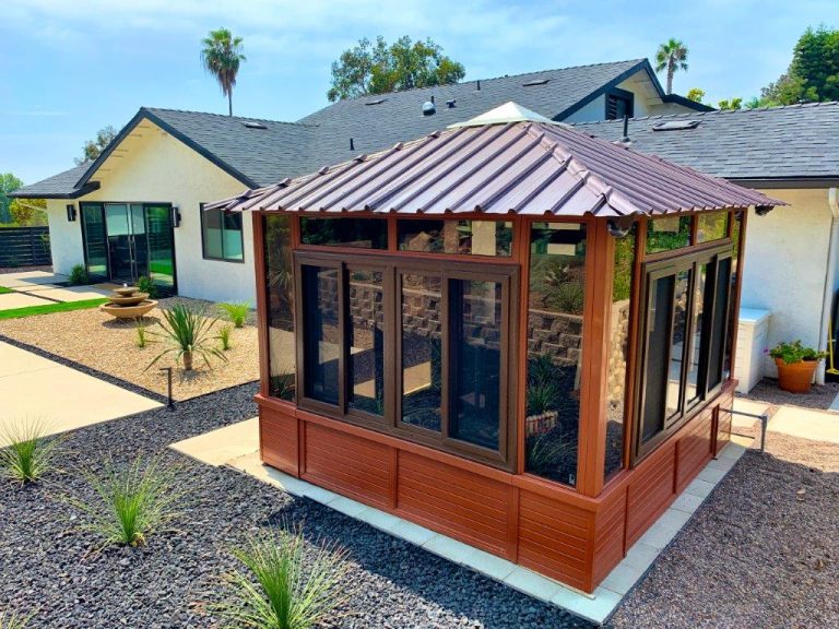 A small glass-and-wood gazebo with a metal roof in a landscaped backyard with desert plants and a modern house.