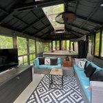 A cozy, enclosed patio with teal sofas, a black rug, a large TV, and a hot tub, surrounded by windows and greenery outside.