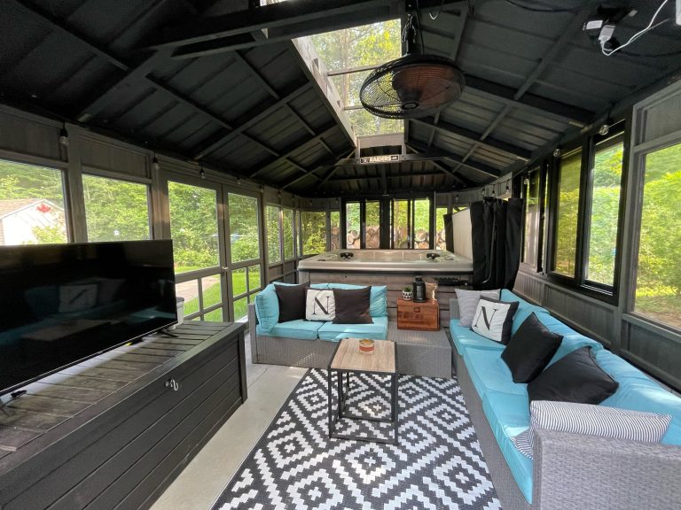 A cozy, enclosed patio with teal sofas, a black rug, a large TV, and a hot tub, surrounded by windows and greenery outside.