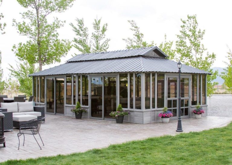 A glass and metal gazebo with a pitched roof, surrounded by potted plants and outdoor seating, set in a green backyard.