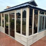A gray, glass-enclosed sunroom with a corrugated roof on a concrete patio.